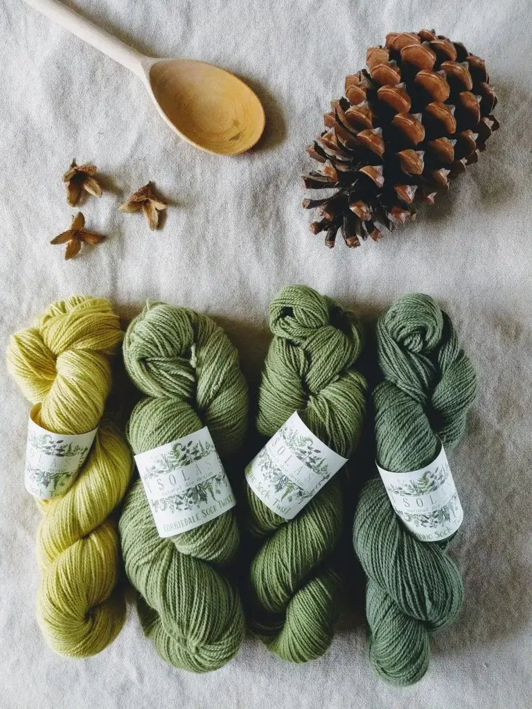 Four skeins of green yarn with labels on a textured surface, accompanied by a wooden spoon and pine cone showcasing the naturally dyed wool used in hand knotted Tradexx kilims